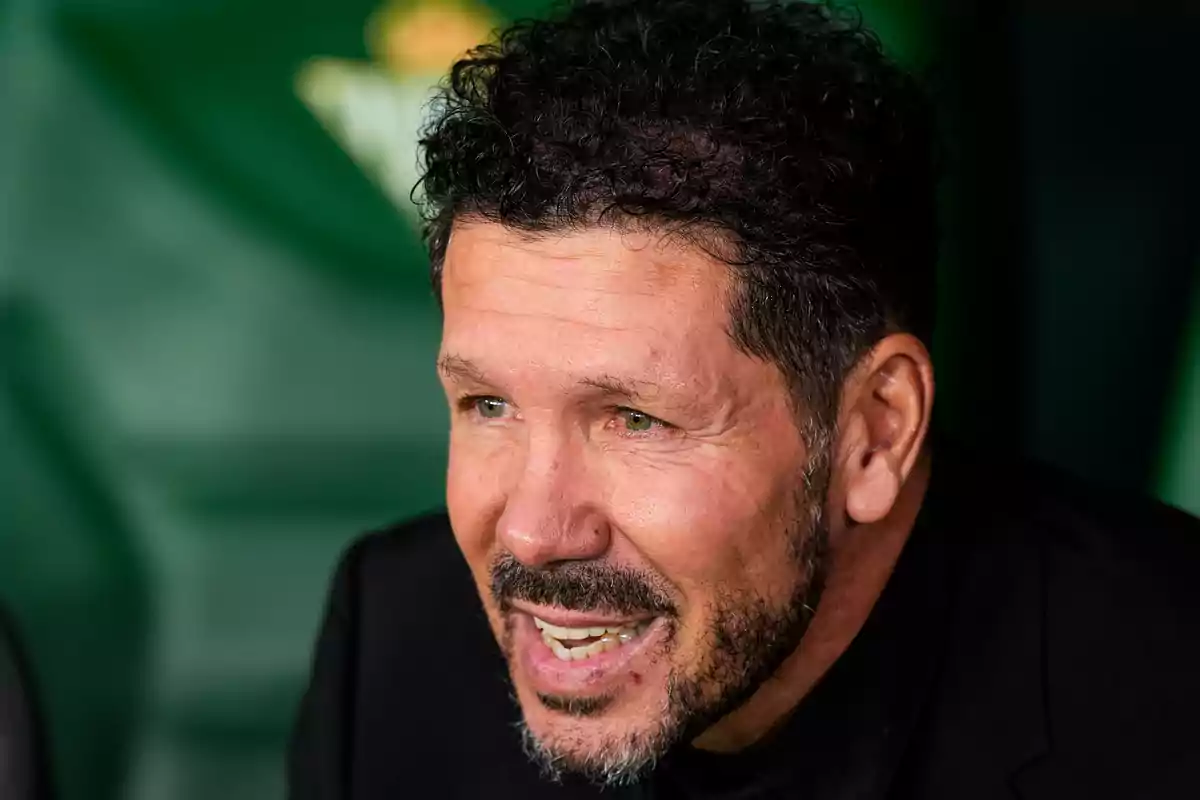 Un hombre con barba y cabello rizado sonríe mientras está sentado frente a un fondo verde.