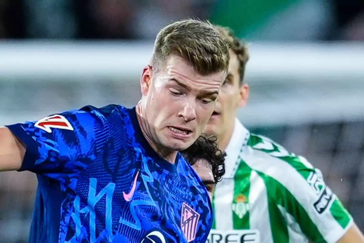 Un jugador de fútbol con uniforme azul compite por el balón con un oponente en uniforme verde y blanco durante un partido.