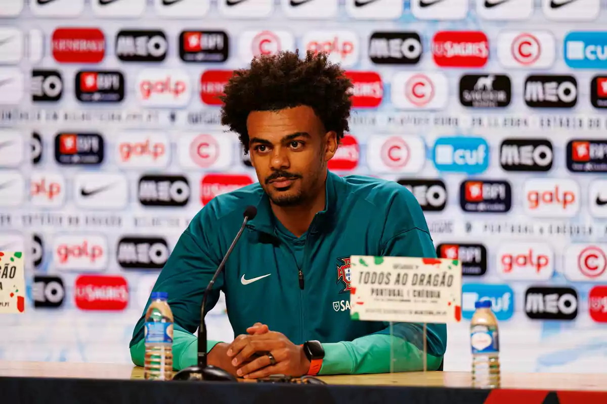 Un hombre con chaqueta verde en una conferencia de prensa con un fondo lleno de logotipos de marcas.