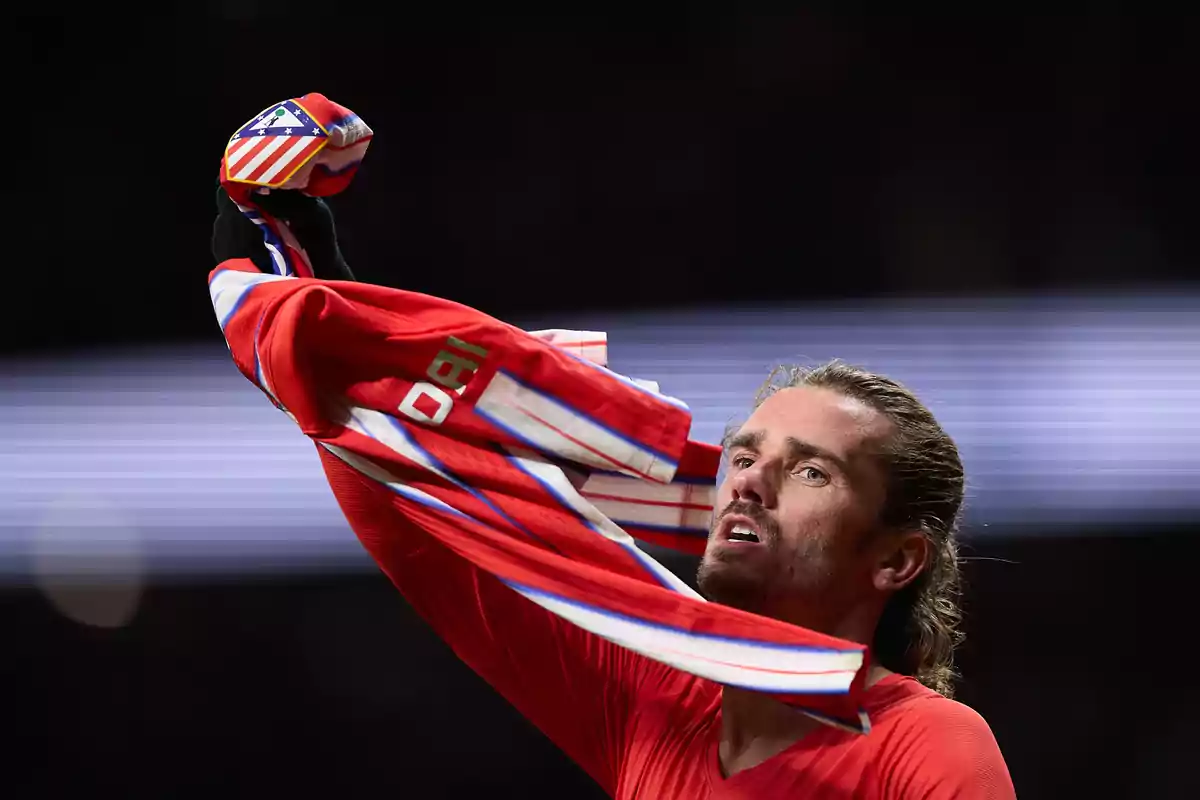 Un jugador de fútbol levanta su camiseta roja con el escudo del Atlético de Madrid en el puño.