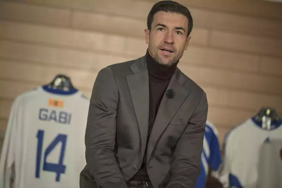 Hombre con saco gris y suéter oscuro sentado frente a una camiseta de fútbol blanca con el nombre Gabi y el número 14