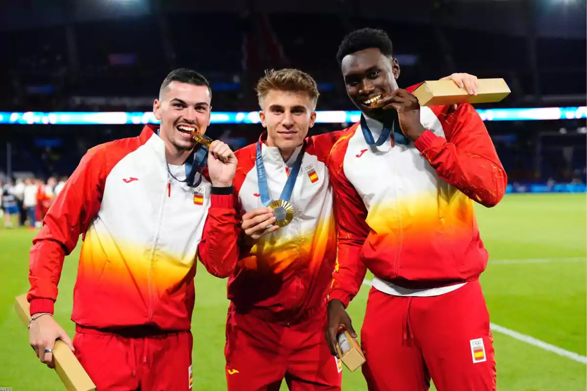 Tres atletas con uniformes de España posan con sus medallas de oro en un estadio, dos de ellos mordiendo sus medallas.