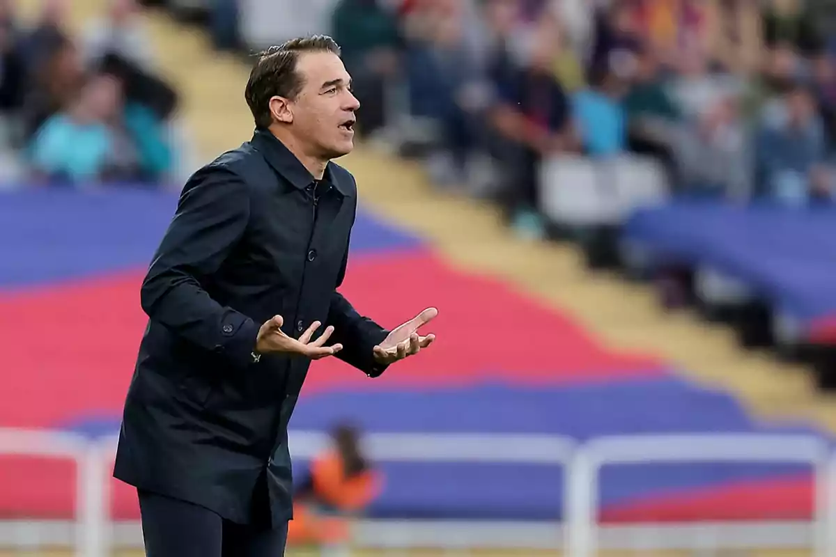 A man in a dark jacket gesturing in a stadium with an out-of-focus audience in the background.