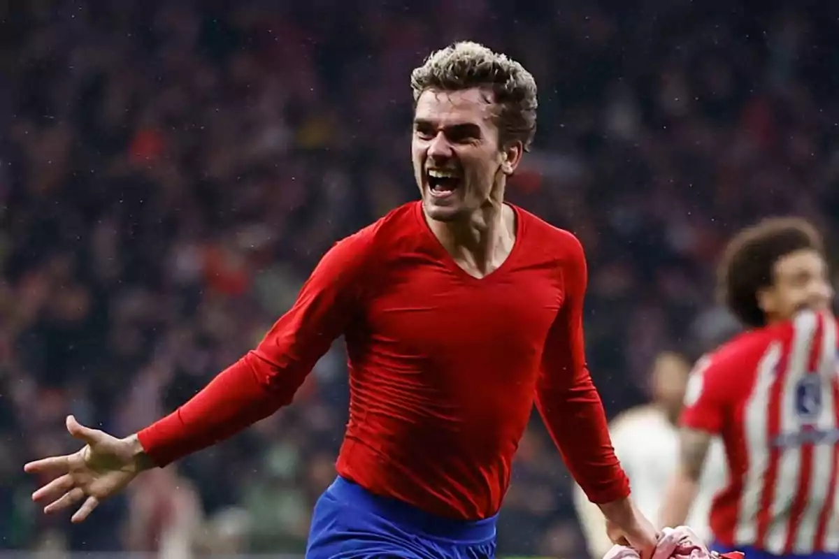 Soccer player celebrating a goal wearing a red jersey and blue shorts in a stadium full of spectators.