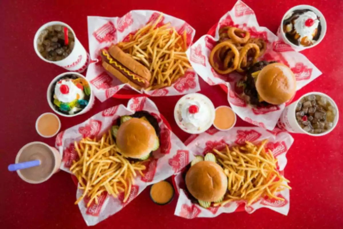 Comida rápida sobre una mesa roja con hamburguesas, papas fritas, aros de cebolla, hot dog, refrescos y postres con crema batida Comida rápida sobre una mesa roja con hamburguesas, papas fritas, aros de cebolla, hot dog, refrescos y postres con crema batida