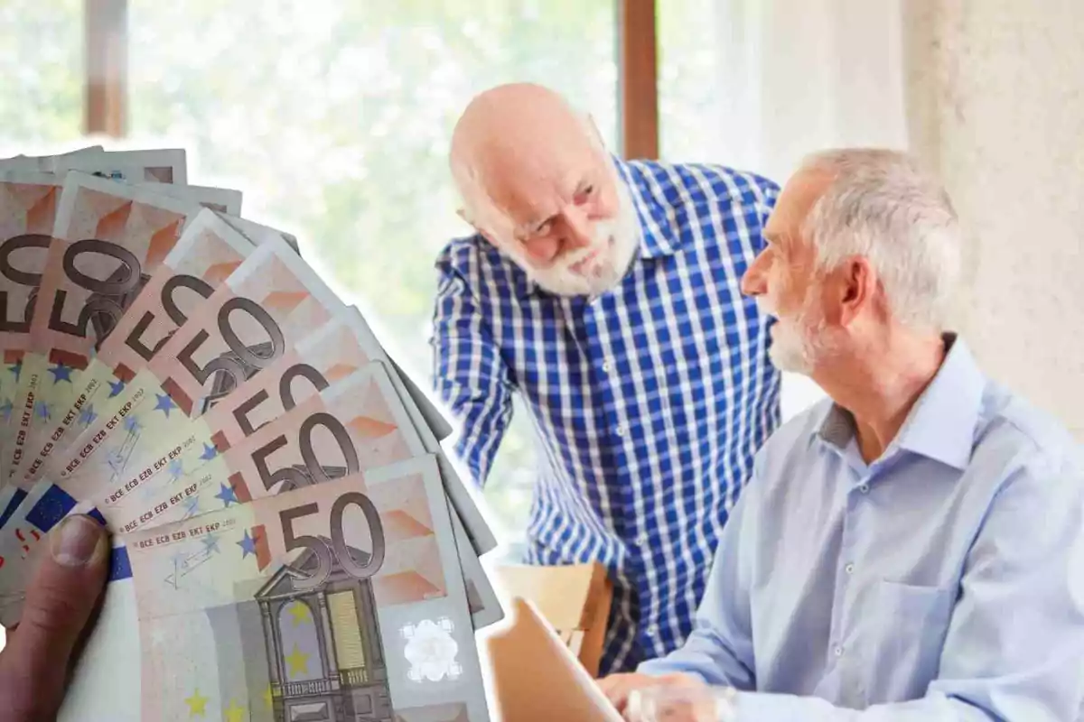 Dos hombres mayores conversan mientras uno de ellos sostiene varios billetes de 50 euros.