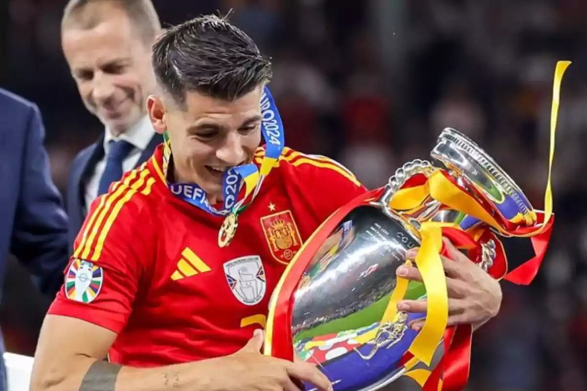 Un jugador de fútbol con la camiseta de España sostiene un trofeo mientras muerde su medalla, con una expresión de alegría en su rostro.