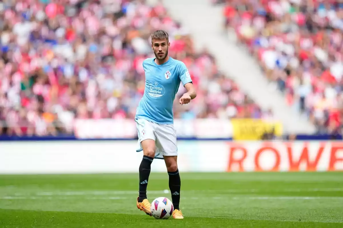 Un jugador de fútbol con uniforme celeste y blanco controla el balón en un estadio lleno de espectadores.