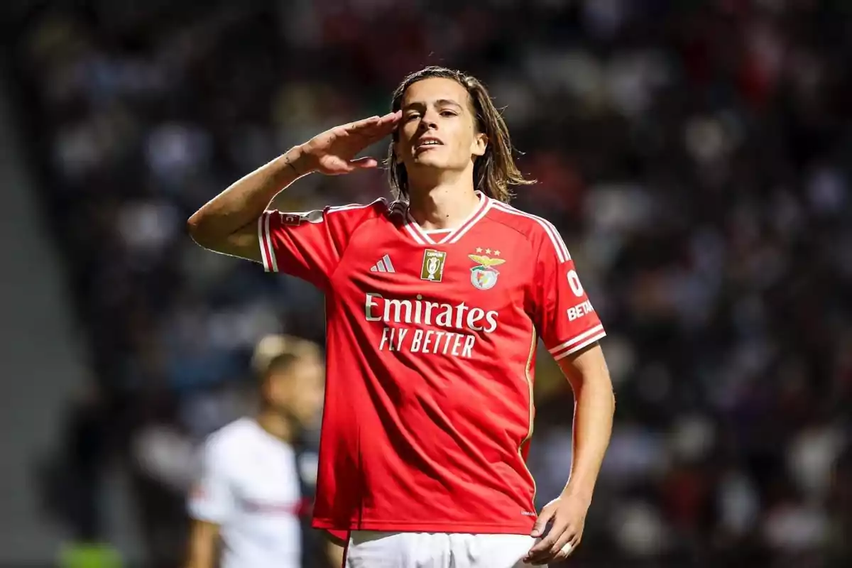 Jugador de fútbol con camiseta roja del Benfica haciendo un gesto de saludo en el campo.