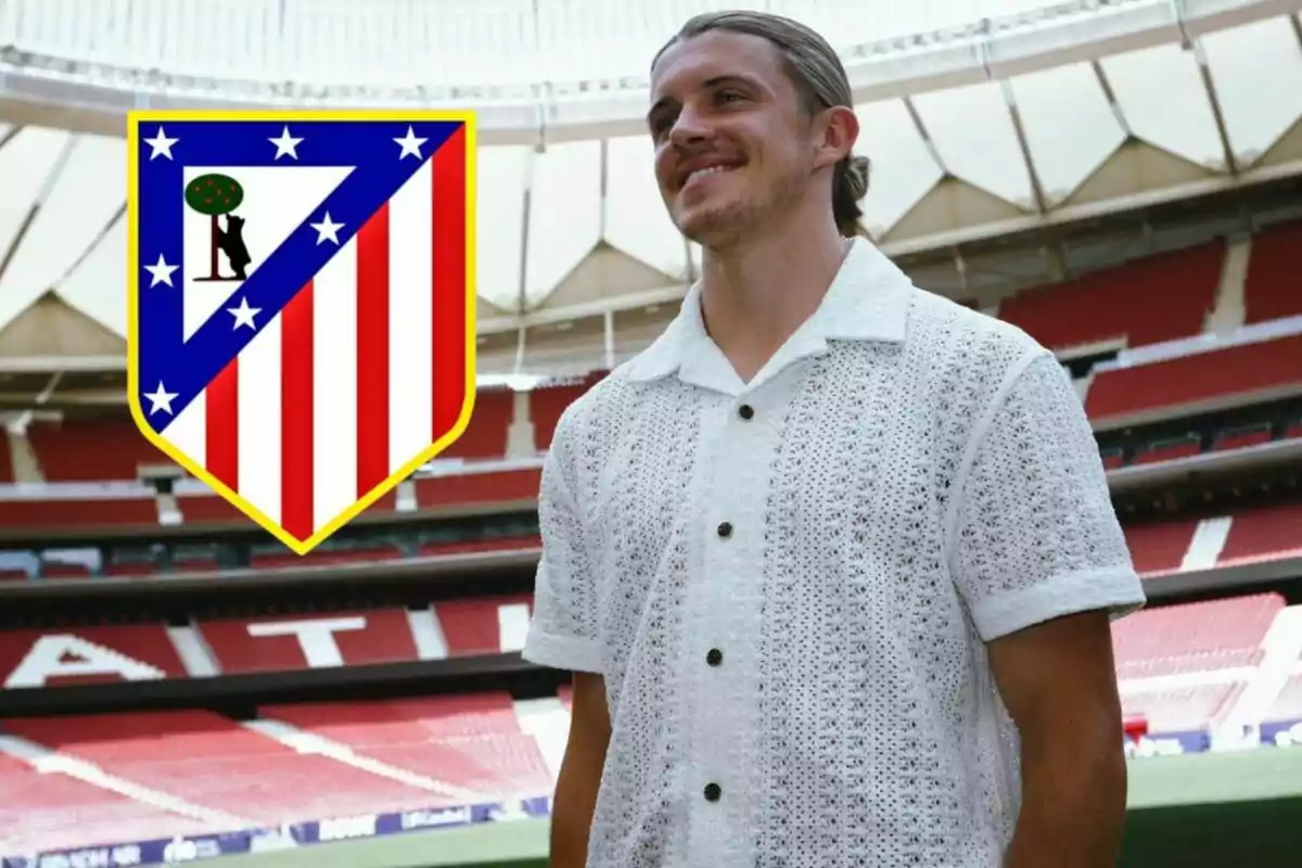 Un hombre sonriente con una camisa blanca de punto posando en un estadio de fútbol con el escudo del Atlético de Madrid a su lado.