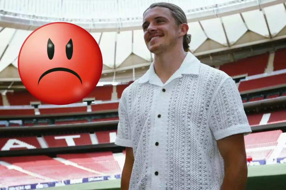 Un hombre sonriente con una camisa blanca de punto está de pie en un estadio de fútbol, mientras un emoji de cara enojada grande se superpone a la imagen.