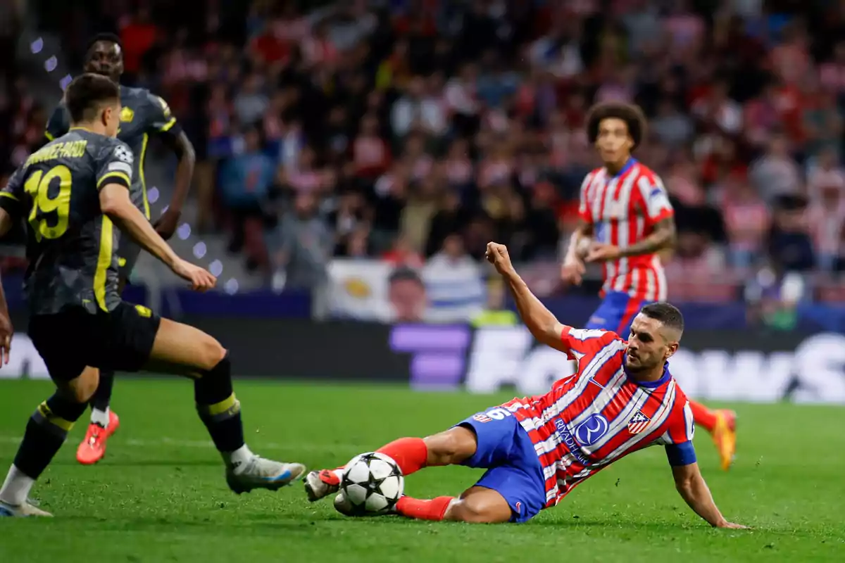 Un jugador de fútbol con uniforme rojo y blanco desliza en el campo mientras un oponente con uniforme gris y amarillo se acerca, con un estadio lleno de espectadores al fondo.