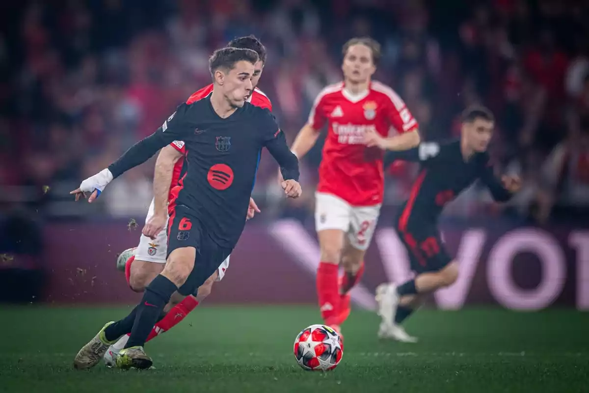 Un jugador de fútbol con uniforme negro conduce el balón en un partido mientras es perseguido por jugadores con uniforme rojo.