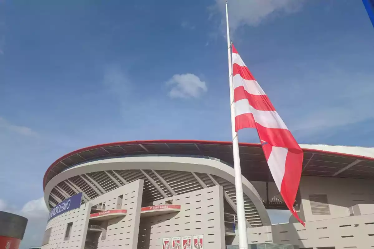 Un estadio moderno con una bandera roja y blanca ondeando a media asta bajo un cielo azul.