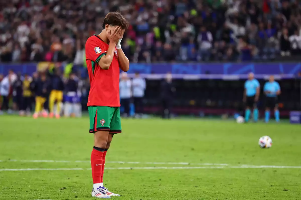 Un jugador de fútbol con el uniforme de Portugal se cubre el rostro con las manos en el campo mientras el público y los árbitros están en el fondo.