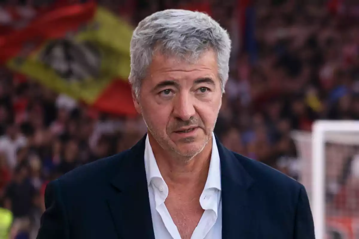 Un hombre de cabello canoso y barba incipiente, vestido con una camisa blanca y un saco oscuro, está de pie frente a una multitud con banderas desenfocadas en el fondo.