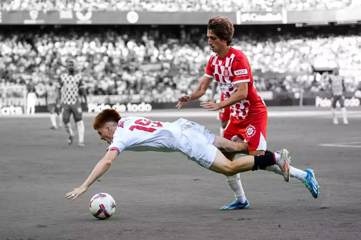 A soccer player in a white uniform falls to the ground while another player in a red uniform follows closely behind him during a game.