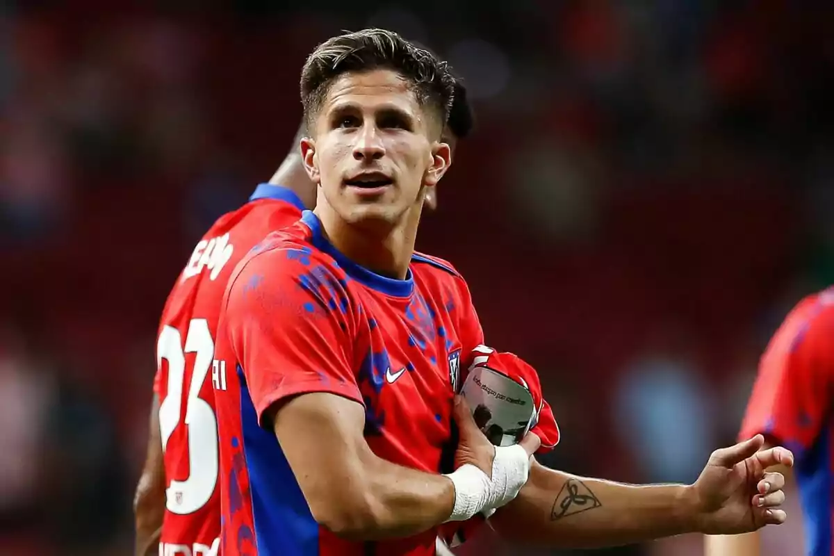 Un jugador de fútbol con uniforme rojo y azul, sosteniendo una camiseta y con un tatuaje en el brazo.