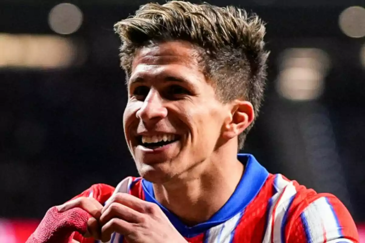 A player smiles while making a heart gesture with his hands, wearing a red and white striped jersey.