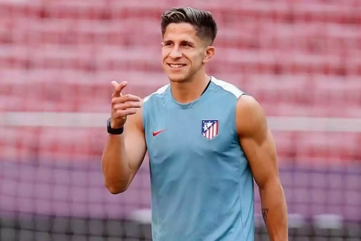 Un jugador de fútbol con una camiseta de entrenamiento del Atlético de Madrid sonríe y señala con la mano.