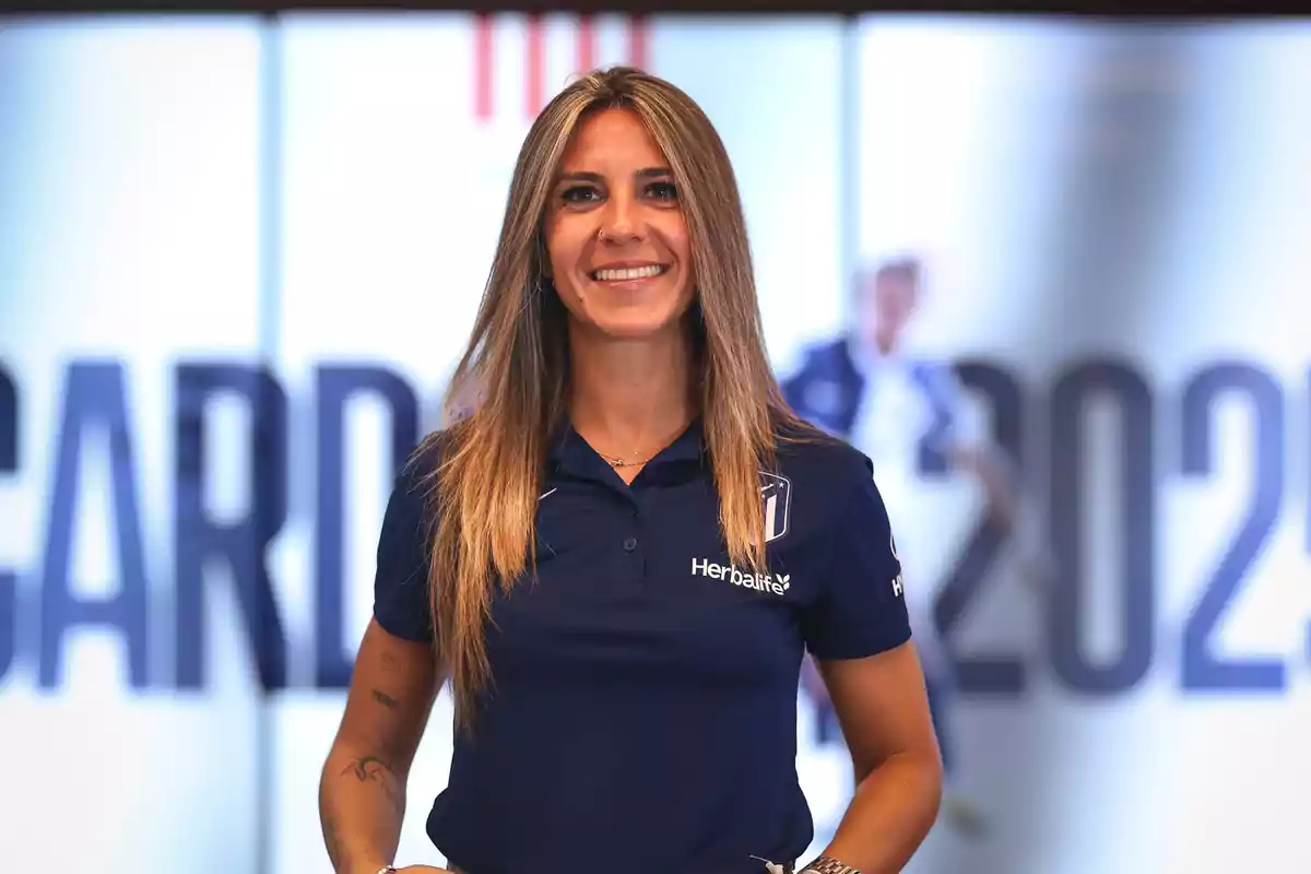 Mujer con camiseta azul de Herbalife sonriendo frente a un fondo borroso con texto.