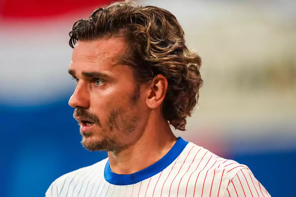 Hombre con camiseta deportiva blanca con rayas rojas y cuello azul, de perfil, con cabello rizado y barba, en un entorno deportivo.