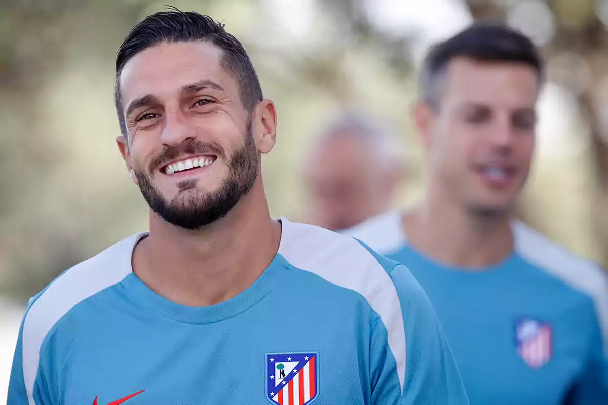 Un hombre con barba y camiseta azul del Atlético de Madrid sonríe a la cámara mientras otra persona desenfocada está detrás de él.