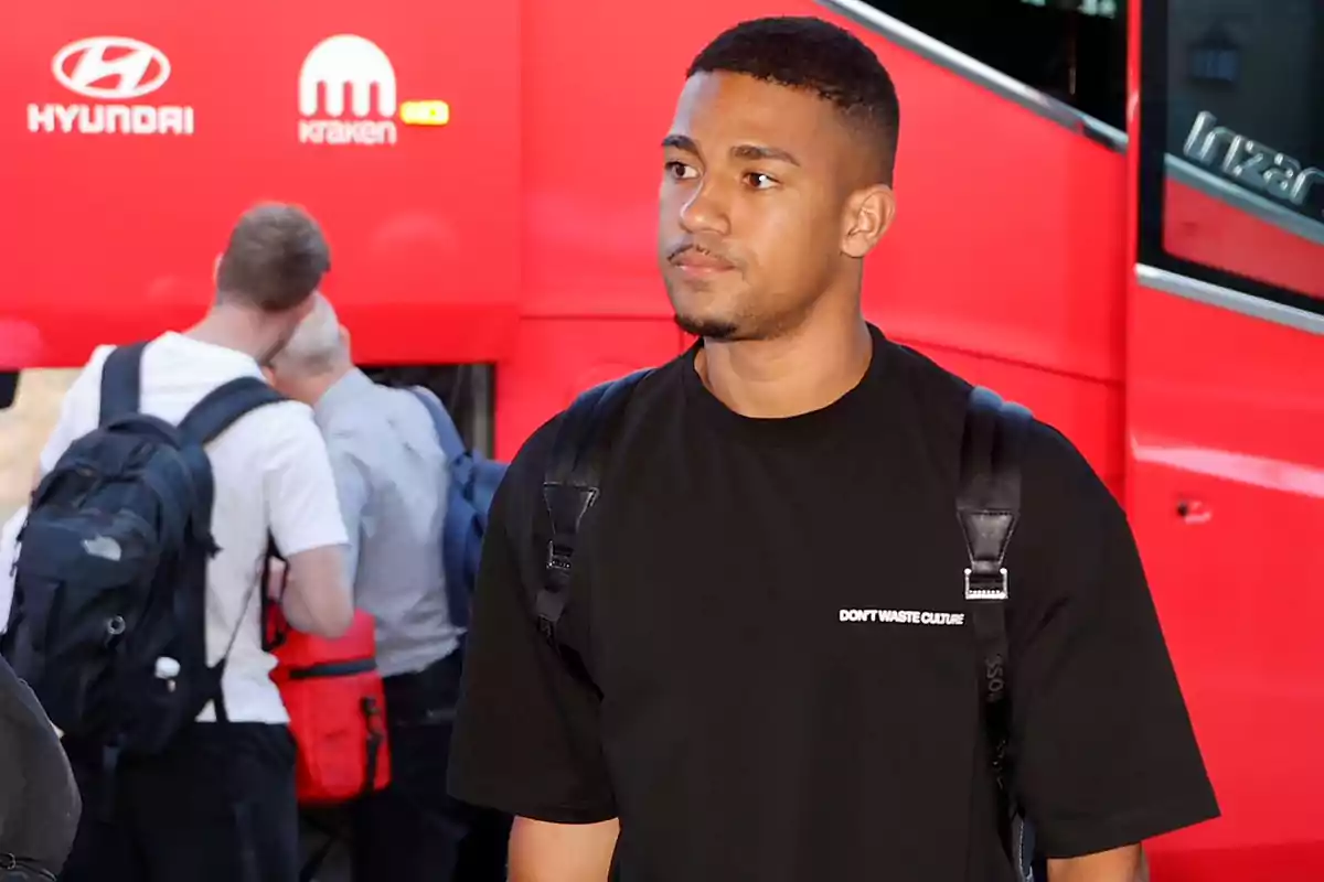 Un hombre con una camiseta negra y una mochila está de pie frente a un autobús rojo con logotipos de Hyundai y Kraken.