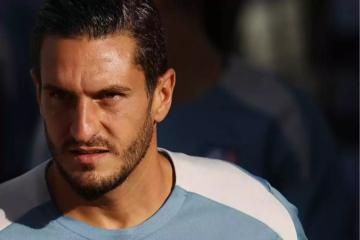 Hombre con barba y cabello oscuro, vistiendo una camiseta azul claro, mirando hacia adelante con expresión seria.