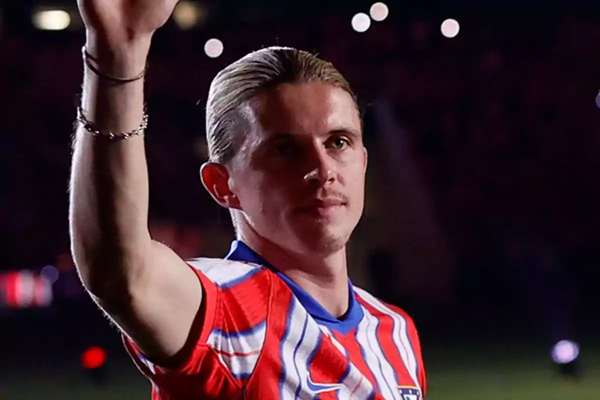 Un jugador de fútbol con uniforme rojo y blanco levanta la mano en un estadio iluminado.