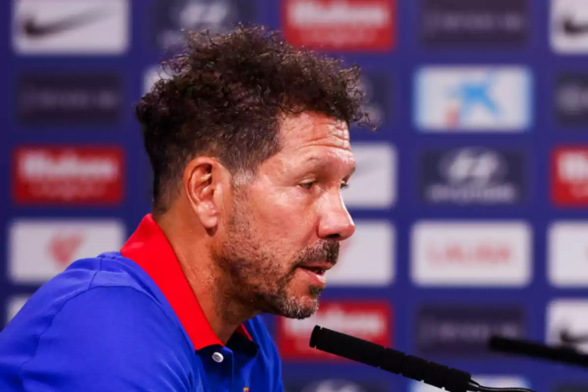 Un hombre con barba y cabello rizado habla en una conferencia de prensa, con un fondo de logotipos de patrocinadores.