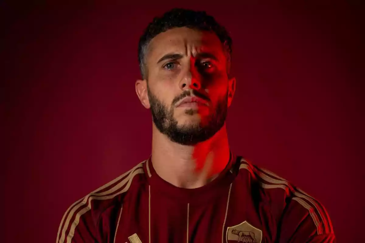 Hombre con barba y cabello rizado, vistiendo una camiseta deportiva roja con detalles dorados, iluminado con luz roja sobre un fondo oscuro.