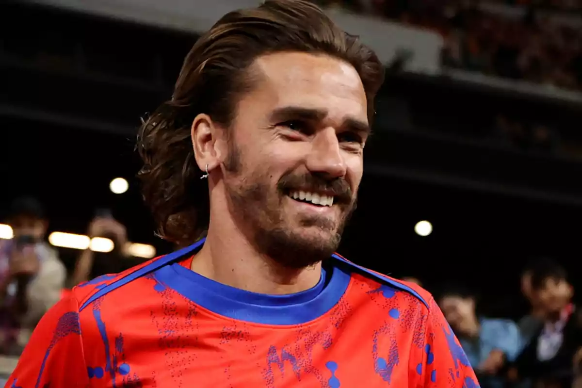 Un hombre con barba y cabello largo sonríe mientras usa una camiseta roja con detalles azules.