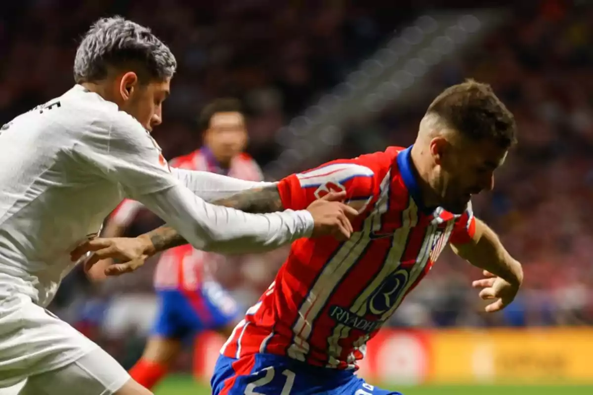 Dos jugadores de fútbol compiten por el balón durante un partido, uno con uniforme blanco y el otro con uniforme rojo y blanco.