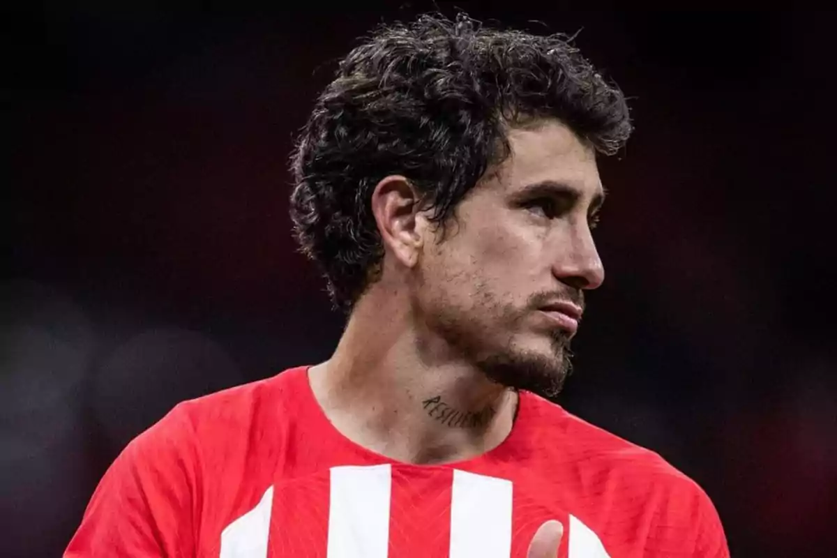 Un hombre con cabello rizado y barba corta, vistiendo una camiseta roja con rayas blancas, mira hacia la derecha. Tiene un tatuaje en el cuello que dice "Resiliencia". El fondo es oscuro y desenfocado.