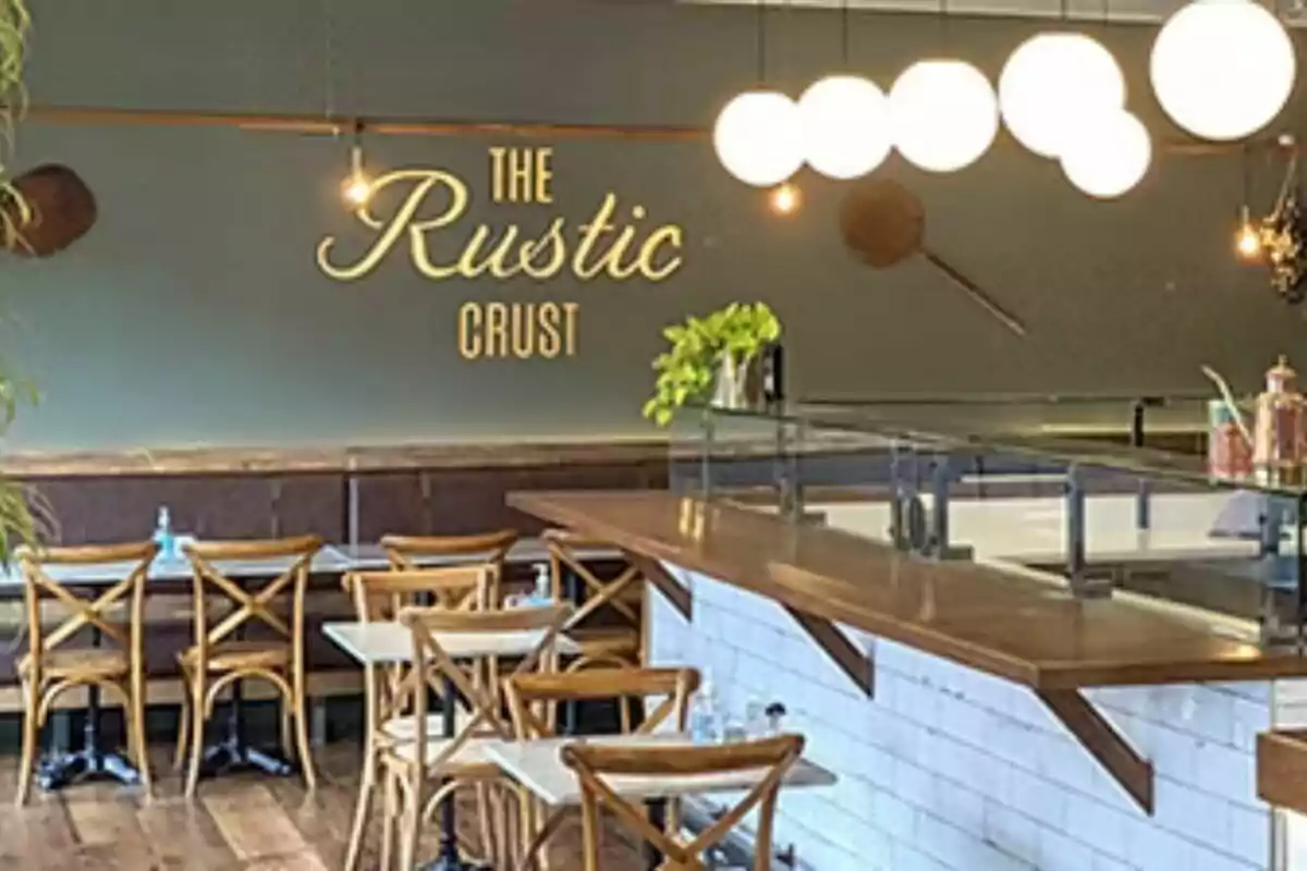 Interior de una cafetería con decoración rústica, mesas y sillas de madera, barra de madera clara, lámparas esféricas colgantes y un letrero en la pared que dice The Rustic Crust
