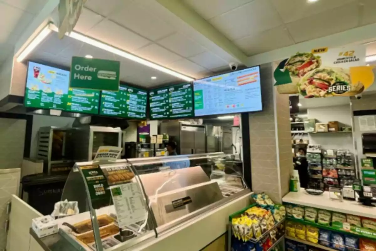 La imagen muestra el interior de un restaurante de comida rápida -que es un Subway- con un mostrador de pedidos, menús digitales en la parte superior y una variedad de productos en exhibición, como papas fritas y bebidas. La imagen muestra el interior de un restaurante de comida rápida -que es un Subway- con un mostrador de pedidos, menús digitales en la parte superior y una variedad de productos en exhibición, como papas fritas y bebidas.