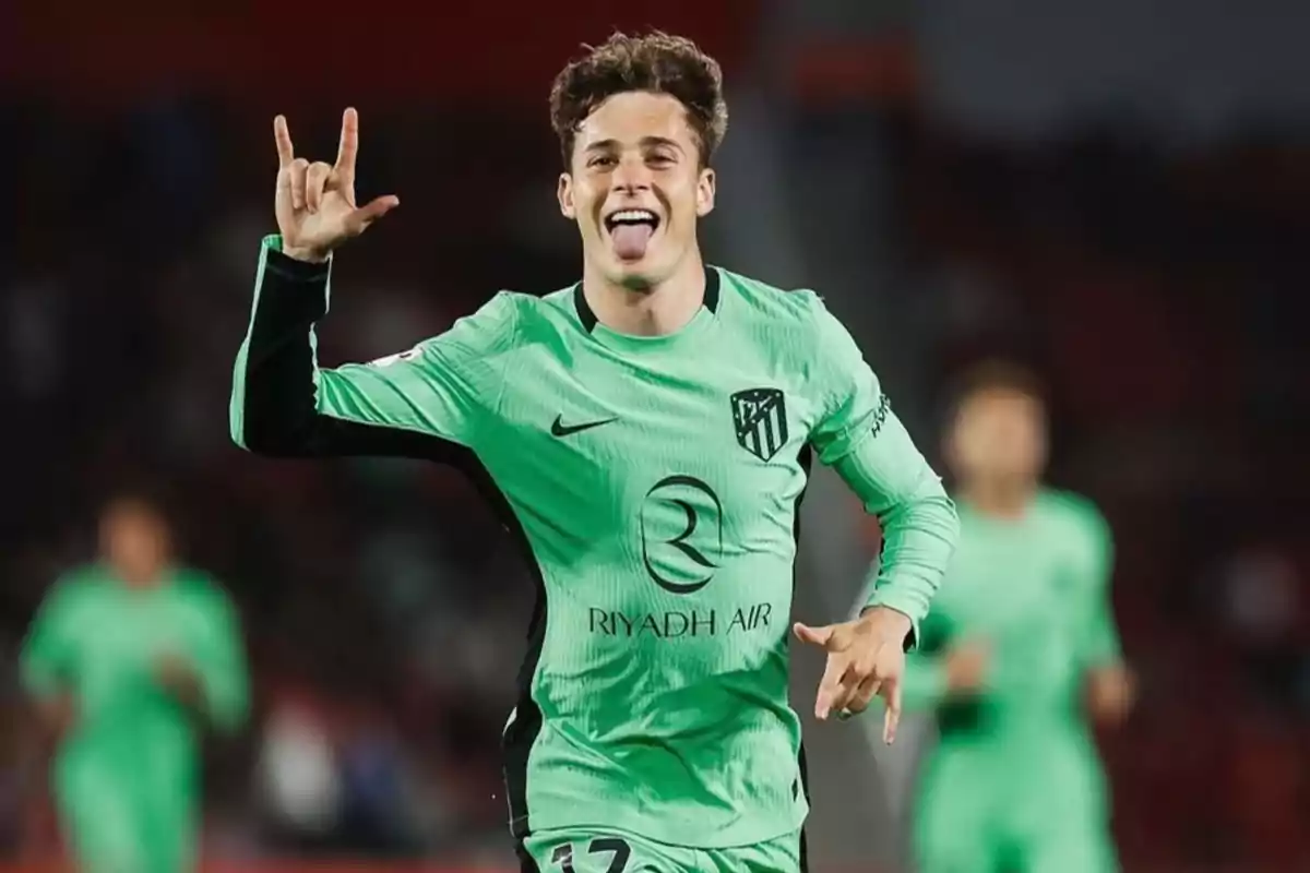 Jugador de fútbol con uniforme verde celebrando en el campo con la lengua afuera y haciendo un gesto con la mano.