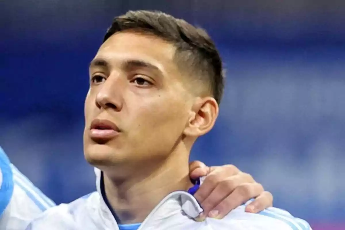 Un hombre con chaqueta deportiva blanca y azul, con una mano apoyada en su hombro, mirando hacia adelante.