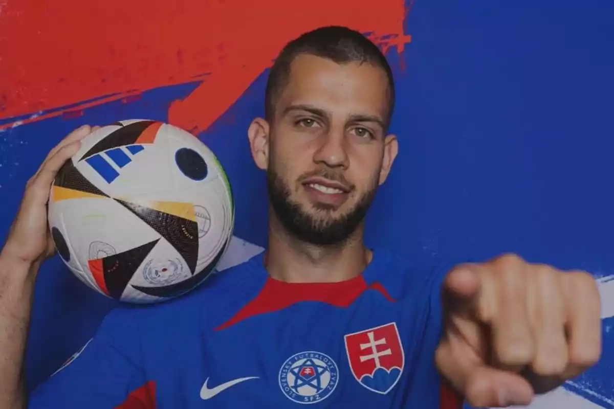 Un jugador de fútbol con uniforme azul y rojo sostiene un balón de fútbol y señala hacia la cámara.