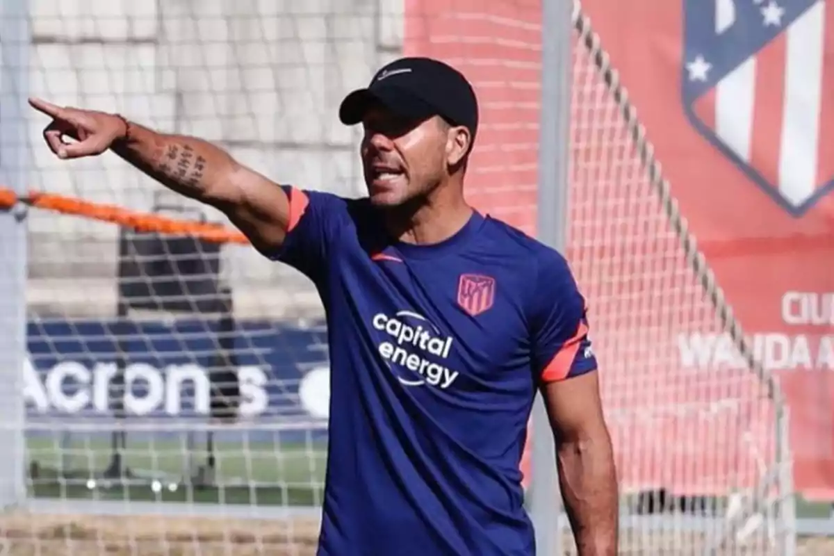 Un entrenador de fútbol con gorra negra y camiseta azul del Atlético de Madrid, señalando con el brazo extendido en un campo de entrenamiento.