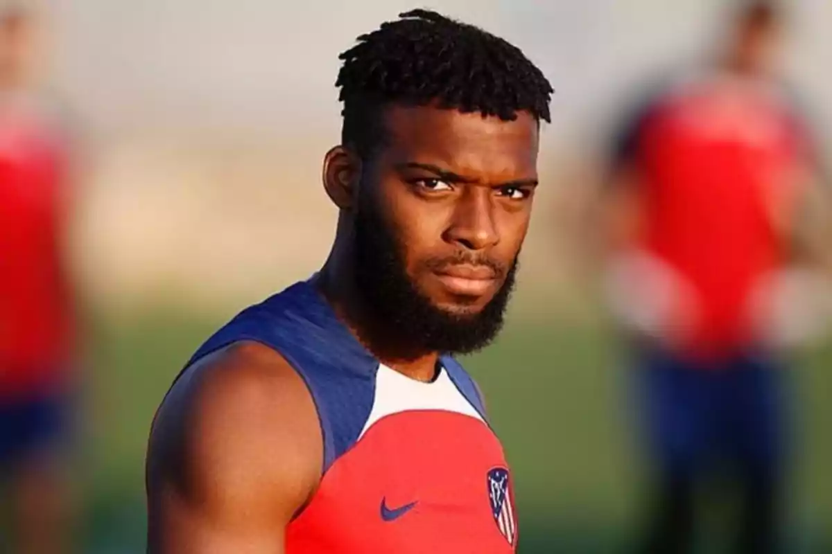 Un jugador de fútbol con barba y camiseta de entrenamiento roja y azul del Atlético de Madrid mira hacia la cámara.