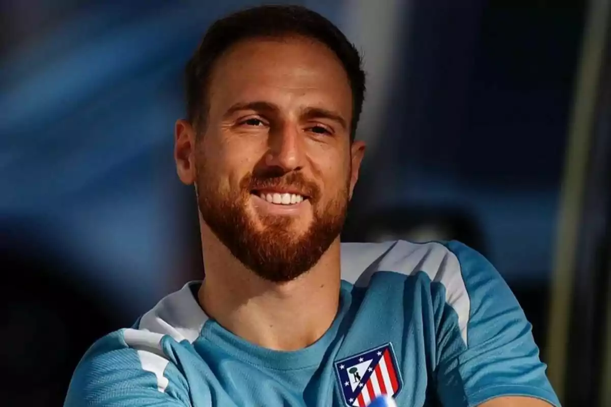 Un hombre con barba y cabello corto sonríe mientras lleva una camiseta deportiva azul con el escudo del Atlético de Madrid.