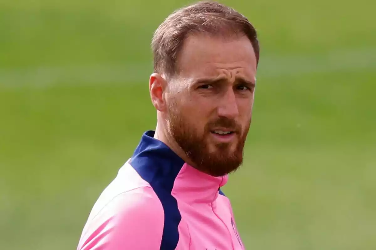 Un hombre con barba y cabello corto lleva una chaqueta deportiva rosa con detalles en azul, mientras está al aire libre con un fondo verde.