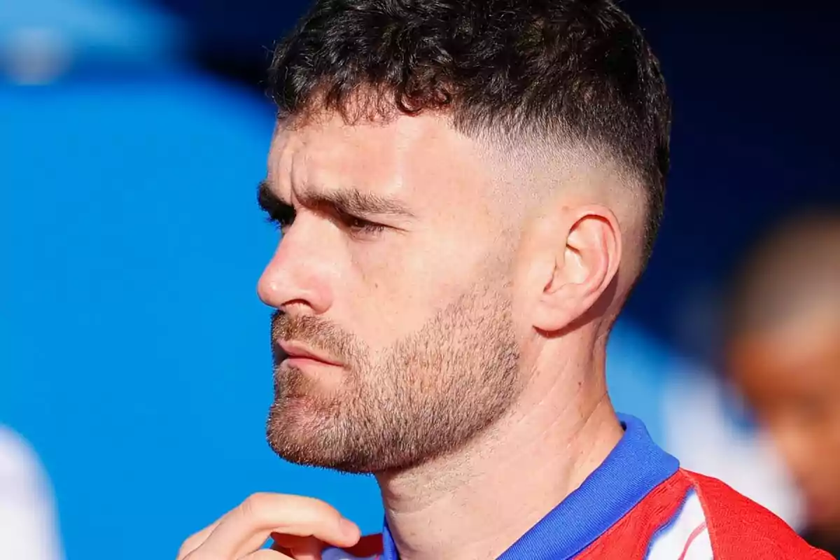 A man with a beard and short hair looks to the left with a thoughtful expression, wearing a red jersey with blue details.