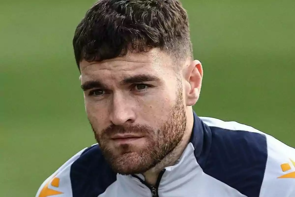 Hombre con barba y cabello corto, vistiendo una chaqueta deportiva, con expresión seria y fondo verde.