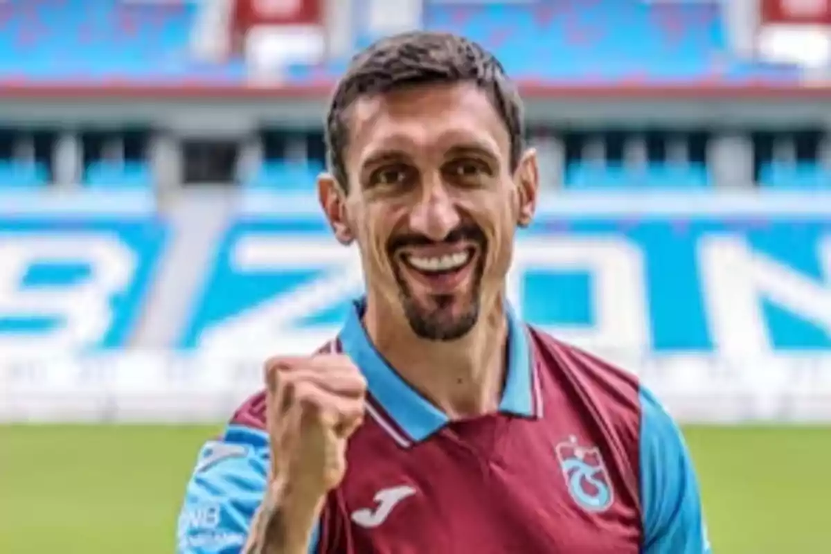 Un jugador de fútbol sonriente con uniforme granate y azul en un estadio.