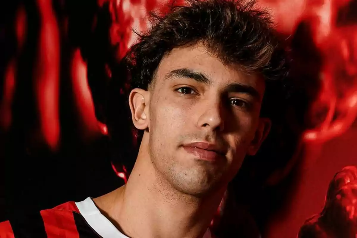 Un hombre joven con cabello oscuro y rizado lleva una camiseta de rayas rojas y negras, posando frente a un fondo rojo.