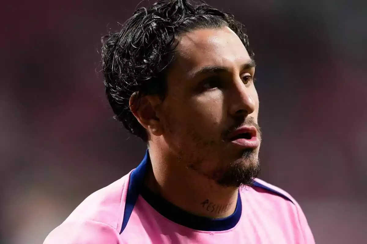 Un jugador de fútbol con camiseta rosa y barba corta mira hacia adelante en un estadio.