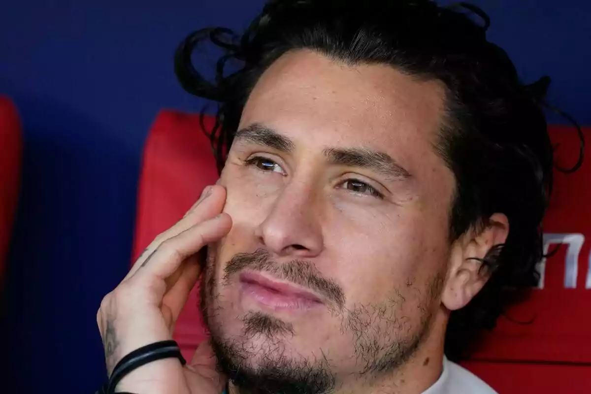 Un hombre con barba y cabello oscuro está sentado, apoyando su rostro en su mano, con una expresión pensativa.